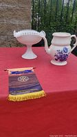 Photo showing a white glass jug with blue floral designs and a white footed pedestal bowl with scalloped edges and decorative cut-outs on a red surface with a Rotary Scarborough Ontario banner nearby.