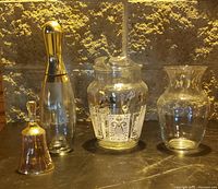 Photo showing four glass items on a counter: a glass bell, a bottle with gold stopper, an etched glass jar, and a clear glass vase.
