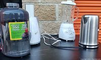 Photo showing two juicers, one blender, and one stainless steel electric kettle placed on a table against a brick wall background.