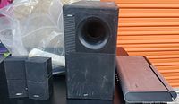 Photo showing four Bose Lifestyle system components: a large black subwoofer with front port, two smaller bookshelf speakers, and a slim rectangular receiver. Components show wear and scratches.