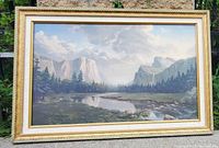 Frontal photo of a framed landscape painting showing mountains, pine trees, river reflecting sky and clouds, set in a decorative gold-tone frame.
