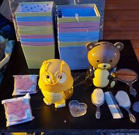 Yellow owl piggy bank, brown bear piggy bank, multiple decorative gift boxes, vintage hair brush and comb set, hand mirror, small containers, and packaged items displayed on a table.