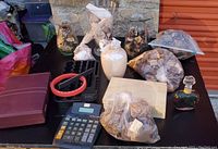 Photo of lot showing four glass vases filled with potpourri, two additional containers including a white ceramic vase and glass decanter, several bags of dried potpourri, a dark box, a black tray, a red circular item, and a large calculator.