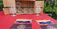 Six Bellevieu Kiek glasses arranged in a cardboard box with two glasses outside, placed on a red tablecloth with two Rotary club pennants.