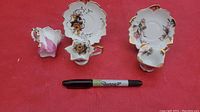Set of three ornate cups and two matching scalloped saucers displayed with Sharpie pen for scale, each piece featuring floral decorations and gold trim.