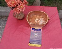 Image showing the ceramic vase with faux flowers and the ceramic bowl placed on a red cloth outdoors, also includes a Rotary club ribbon.