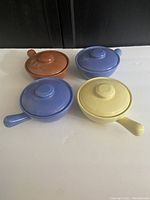 Four Medicine Hat Potteries covered soup bowls with handles and lids in varying colors (yellow, light blue, darker blue, brown) arranged in a square formation on a surface.