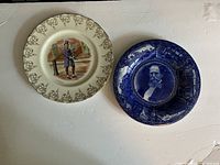Photo of two collectible plates: left plate with Mr. Micawber on a London street with grapevine decorative rim, right plate with blue and white portrait of Charles Dickens surrounded by scenes.