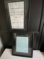 Photo of two black framed items propped against black wall. Top frame contains crew call sheet with signatures. Bottom frame contains blue script cover sheet with signatures.