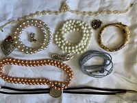 Photo of assorted costume jewelry items including multiple necklaces (pearl bead and pop-it bead styles), bracelets, brooch, and small broken figurine.