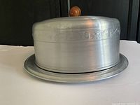 Side view of vintage aluminum cake cover and plate with wooden acorn knob, showing engraved oak leaves pattern.