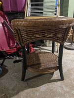 Front angle view of the round wicker side table showing woven rattan top and lower shelf supported by black wooden legs.