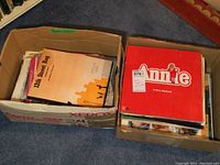 Two boxes on floor, one shows music sheets and 42nd Street Rag music sheet visible, other box shows red Annie musical vinyl album on top with many other albums inside.