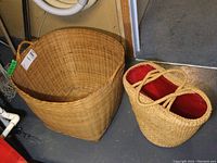 Two baskets side by side on floor. Wicker laundry basket is large and rectangular. Straw shopper tote is oval with red lining and braided handles.