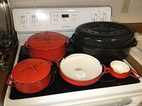 All pieces arranged on stove: black speckled roasting pan, three red lidded pots, shallow pan, saucepan and gravy container