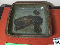 Top and angled views showing green and brown glazed surface with artistic abstract pattern and four cigarette rests.