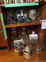 Shelf showing bird paperweights, decorative mask, ceramic jug with dragon, beer mugs, decanters, figurines, and candle.