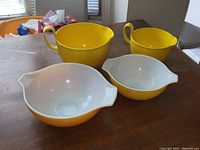 Four mixing bowls on wooden table: two large Pyrex daisy patterned bowls in foreground, two bright yellow Dansk Gourmet design mixing bowls with handles and spouts in background.