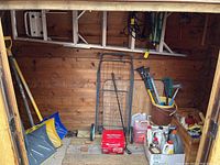 Photo showing three snow shovels, metal shopping buggy, battery charger, and various containers including solvents and watering equipment inside a wooden shed.