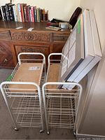 Two white rolling wire storage carts with wheels, placed side by side with two white boxed photo frames resting against one cart