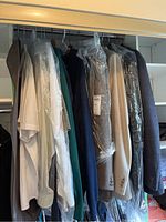 Multiple men's jackets, shirts, and sweaters hanging in a closet rack, some covered in protective dry-cleaning plastic bags. Includes light and dark coloured items with varied fabrics and styles.