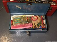 Top view of one blue metal toolbox open showing art materials like colored pencils, a pink comb, and small containers.