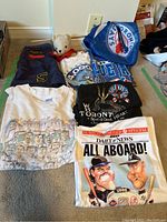 Photo showing folded vintage t-shirts including Toronto Blue Jays and Who concert shirts, Blue Jays cap and flag, and navy shorts with yellow S emblem.
