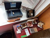Overall view of all items displayed on table and window ledge, showing watches in black box, assorted cufflinks, copper cuff, utility cutter with sheath, vintage case, and jewelry box.