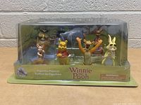 Photo of the sealed Disney Winnie the Pooh figurine playset featuring six characters: Winnie the Pooh, Tigger, Rabbit, Owl, Eeyore, and Kanga. Clear front packaging and green cardboard backer with branding.
