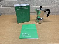 Polished aluminum stovetop espresso maker, original green box, and instruction manual displayed on a flat surface against a white brick background.