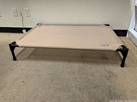 Full view of the K & H dog bed showing the tan mesh fabric stretched over a black metal frame with four legs on a carpeted floor.