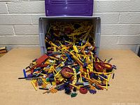 Large plastic tub filled overflowing with assorted K'nex rods, connectors, wheels, and gears in various colors on a wood table against a brick wall.
