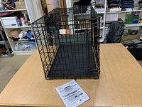 Folded black metal wire pet crate with plastic tray base and instruction sheet on table surface