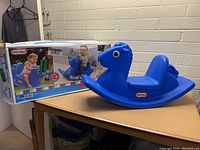 Blue plastic rocking horse on a wood surface next to its packaging box showing a child playing on the toy.