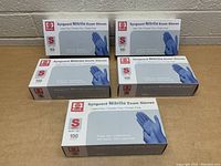 Five boxes of Synguard Nitrile Exam Gloves arranged on a table against a cinderblock wall, each box labeled small size with 100 gloves per box.