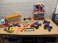 Overview of all kids tool toys arranged on table showing variety including boxed tool set, saws, drills, vacuum and clamps.