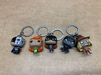 Five Pop figure keychains of Batman with cyborg face, a red-haired woman, black-clad figure, gray cat, and scarecrow arranged on a wooden surface.