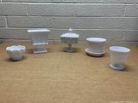 Front view of five vintage milk glass pieces displayed on a wooden table against a brick wall background.