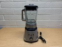 Front view of Cuisinart blender on tan surface against brick wall, showing glass jar with lid and power cord beside base.