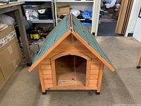Front angled view of the wooden dog house showing green shingled roof and wooden panels.
