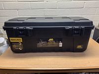 Front view of black Plano sportsman's trunk showing heavy-duty label, dual latches, and brand label in center. Trunk closed with visible molded handle details on lid.