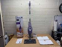 Shark Steam Pocket Mop standing next to original box, instruction manual, two white mop pads, and measuring cup on a wooden surface with brick wall background.