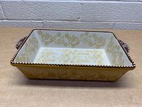 Ceramic rectangular baking dish with yellow floral vines inside and outside, brown decorative rim, white detailing on handles, top-down angle.