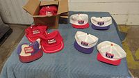 Box containing red GMC Trucks snapback hats and a collection of trucker hats displayed on a blue padded surface. Hats include various red and purple cap styles with embroidered GMC logos and Interstate highway numbers.
