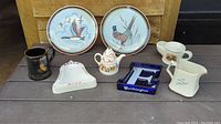 Photo showing all 8 vintage pottery items including two Stangl chop plates, two whiskey advertising ashtrays, Wade Christmas teapot, Wade John Begg pitcher, and two mugs.