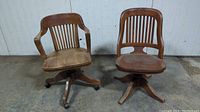 Front view of two antique oak banker chairs with vertical slat backs. One chair has arms and casters; the other is armless and missing casters.