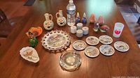 Overview of the entire collection including salt & pepper shakers, souvenir plates, ashtrays, cruets, and figurines arranged on wooden table to show relative sizes compared to a water bottle.