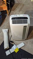Full view of the white DeLonghi portable air conditioner with hose and window vent attachments on the floor.