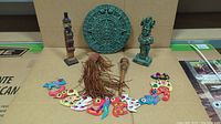 All 6 Mayan collectibles arranged on table with cardboard background showing the full set including the sun calendar, figurines, rattle, and feathered necklace.