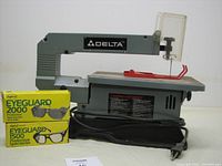 Front view of 13-inch Delta scroll saw with power cord and two yellow boxes of Eyeguard safety goggles placed beside it.
